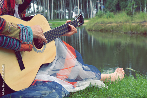 A hipster woman plays the guitar on the grass by the lake. Retrostyle 70s