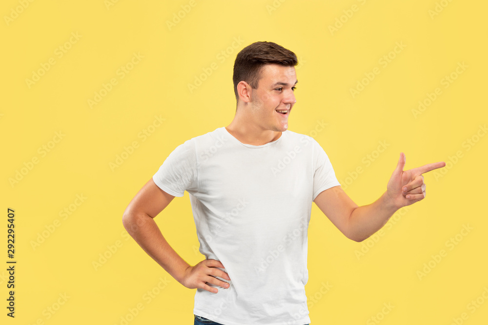 Caucasian young man's half-length portrait on yellow studio background. Beautiful male model in shirt. Concept of human emotions, facial expression, sales, ad. Pointing at side, looks happy.