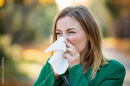 Obraz na plátně Sick young woman with cold and flu standing outdoors, sneezing, wiping nose with handkerchief, coughing