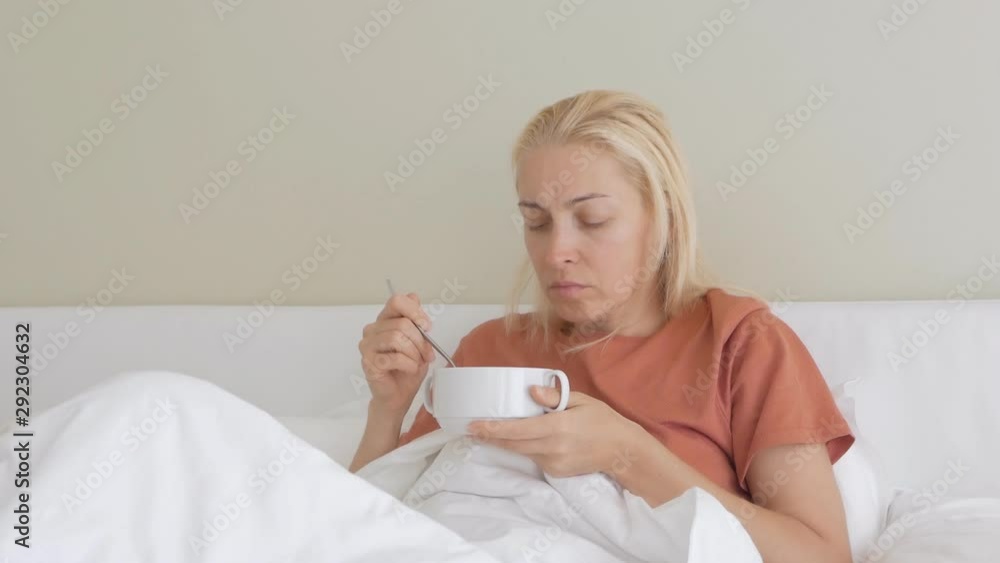 A woman with a sad painful face sitting on bed under white blanket and eating from a plate of soup. A woman with a sad painful face sitting on bed under white blanket and eating from a plate of soup.