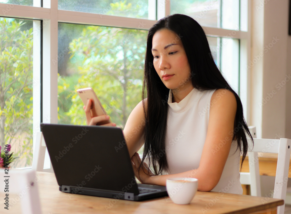 Beautiful Asian business woman sitting alone in the coffee shop with computer laptop on the table , holding mobile phone and reading text on mobile phone attentively. Beautiful Asian business woman sitting alone in the coffee shop with computer laptop on the table , holding mobile phone and reading text on mobile phone attentively.