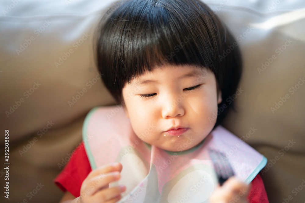Adorable healthy 2 years old Asian toddler enjoys her snack, eating by herself using a hand and fork while looking happy in the living room, selective focus in concept toddler learning to eat food. - obrazy, fototapety, plakaty 