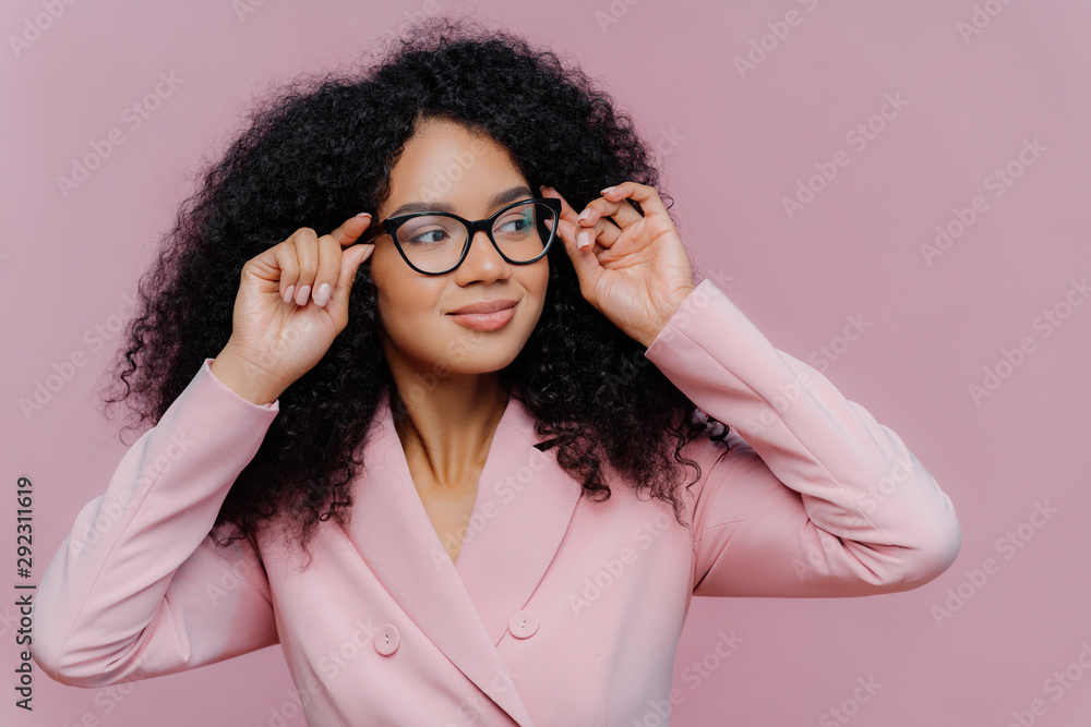Headshot of attentive female boss keeps hands on frame of glasses, looks pensively away, wears rosy formal suit, has curly Afro hairstyle, poses against purple background. People, business, ethnicty - obrazy, fototapety, plakaty 