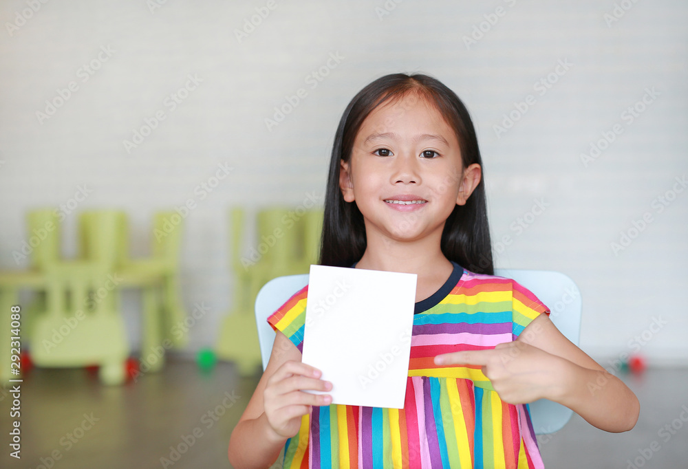 Portrait little smiling Asian child girl holding blank white paper card in her hand with pointing. Kid showing empty paper note copy space in children room. Portrait little smiling Asian child girl holding blank white paper card in her hand with pointing. Kid showing empty paper note copy space in children room.