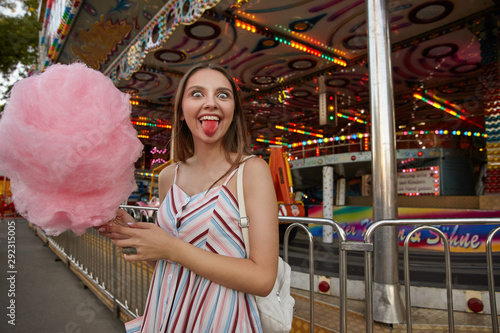 Funny open eyed brunette female with long hair in light summer dress posing over amusement park, looking at camera happily and showing pink tongue after eating cotton candy