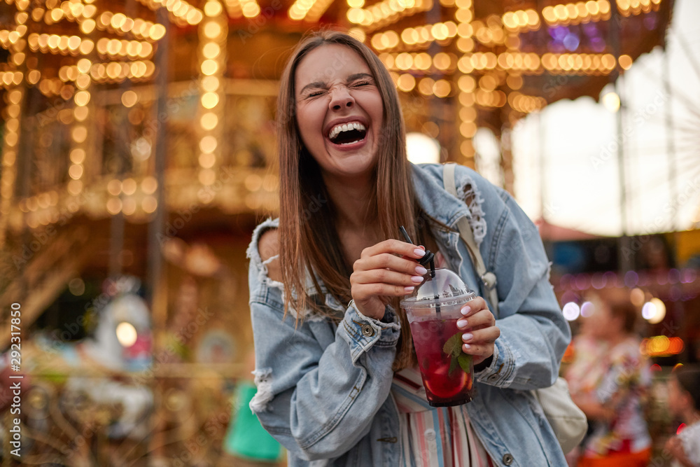 Cheerful beautiful young woman with brown hair in casual clothes drinking lemonade while walking in amusement park, laughing loud with closed eyes and puckering