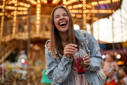 Cheerful beautiful young woman with brown hair in casual clothes drinking lemonade while walking in amusement park, laughing loud with closed eyes and puckering © timtimphoto
