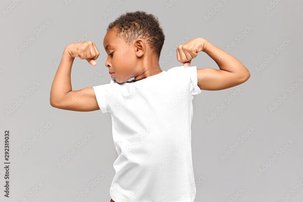 Children, fitness and bodybuilder concept. Studio shot of athletic muscled African American boy in casual t-shirt demonstrating strength by tensing bicep muscles, having proud confident look Children, fitness and bodybuilder concept. Studio shot of athletic muscled African American boy in casual t-shirt demonstrating strength by tensing bicep muscles, having proud confident look