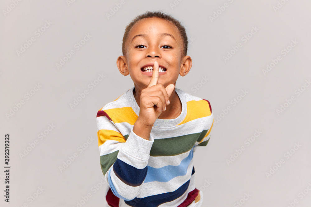 Φωτογραφία Handsome black boy of school age smiling broadly and holding fore finger at his mouth, asking to keep silent because he toddler sister is sleeping