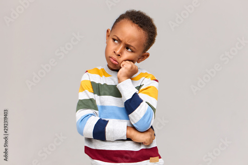 Waist up shot of moody grumpy African schoolboy in sweater holding hand on his face, pouting lips and frowning, sulking with his greedy friend who does not want to share toys. Complain or disagreement photo
