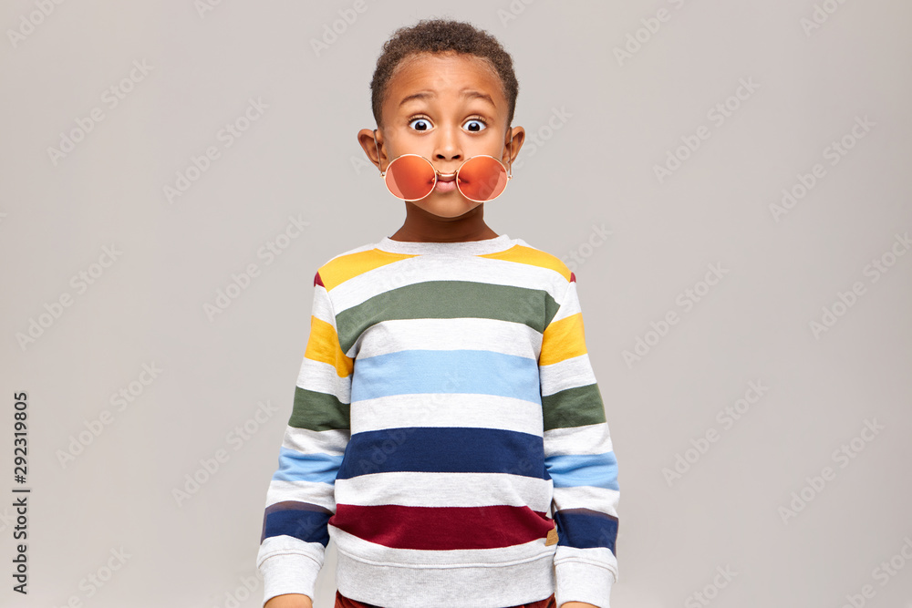 Oops. Funny bug eyed dark skinned little boy in stylish multicolored jumper staring at camera in full disbelief, round pink sunglasses slipped off his eyes. Surprise, shock and astonishment - obrazy, fototapety, plakaty 