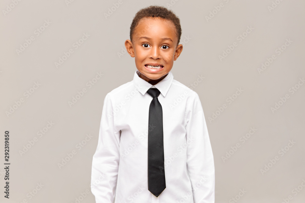 Shy or scared emotional African schoolboy in white shirt and black tie baring his teeth ...