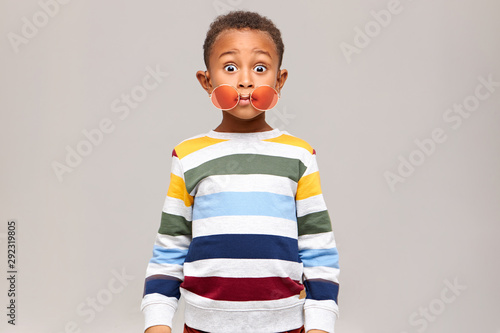Oops. Funny bug eyed dark skinned little boy in stylish multicolored jumper staring at camera in full disbelief, round pink sunglasses slipped off his eyes. Surprise, shock and astonishment