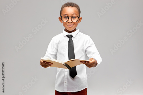 Studio image of funny smart akward African schoolboy in formal clothes and eyeglasses eager to learn, holding open book in his hands and smiling. Enthusiastic dark skinned A-student learning at school photo