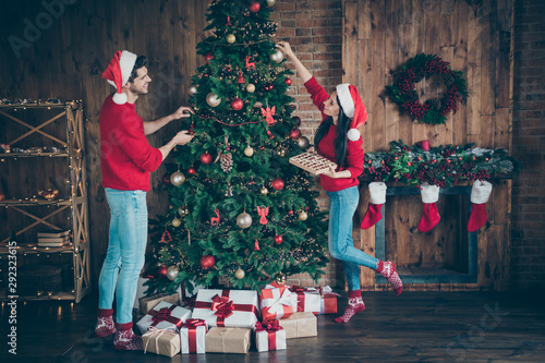 Full size profile side photo of two romantic spouses people holding baubles decorate christmas tree for x-mas wearing cap hat in house full of newyear decoration lights illumination indoors