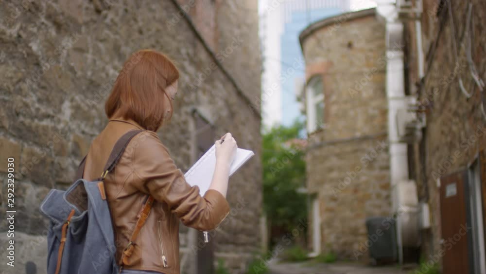 Slowmo tracking with rear view of creative young woman with red hair standing in alleyway and sketching old brick building Slowmo tracking with rear view of creative young woman with red hair standing in alleyway and sketching old brick building
