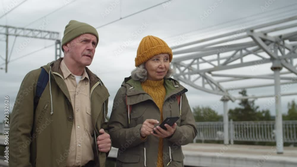 Medium shot of Caucasian senior couple walking along train station, talking and looking at smartphone screen aged woman holding in her hands Medium shot of Caucasian senior couple walking along train station, talking and looking at smartphone screen aged woman holding in her hands