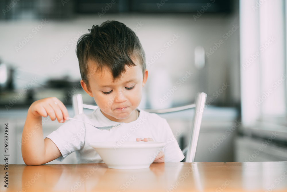 A little boy in the kitchen eating oatmeal from a white plate A little boy in the kitchen eating oatmeal from a white plate