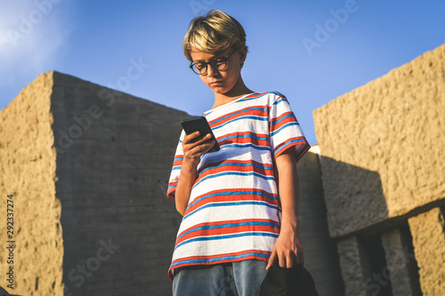 Portrait of a beautiful teen using smartphone social app standing in a urban context. Young trendy boy watching social video online on cellphone. Youth urban lifestyle and new technology concept. photo