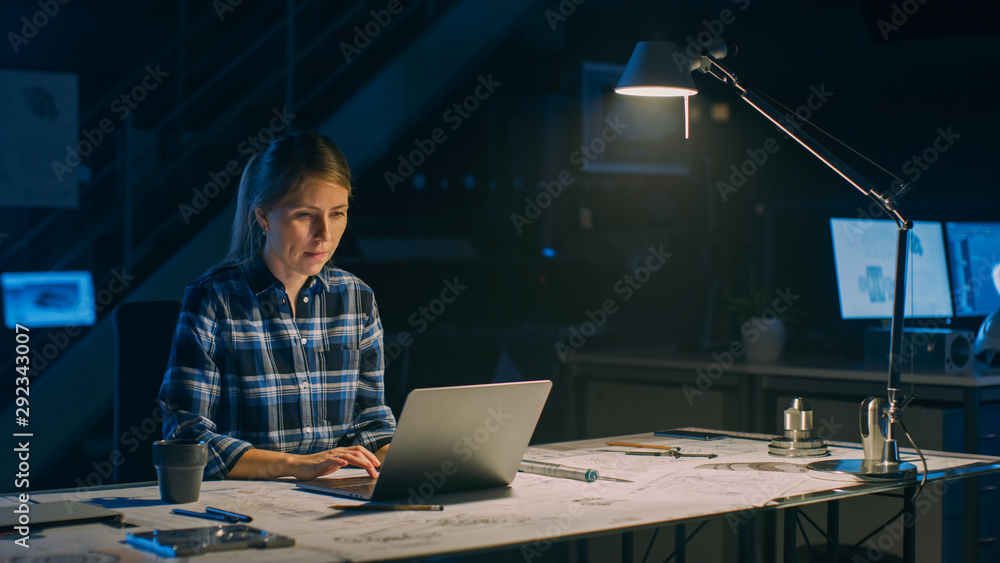 Beautiful Female Engineer Sitting at Her Desk Works on a Laptop Computer. Blueprints Lying on a Table. In the Dark Industrial Design Engineering Facility. Warm Light Lamp