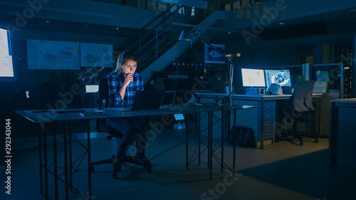 Beautiful Female Engineer Sitting at Her Desk Works on a Laptop Computer. Blueprints Lying on a Table. In the Dark Industrial Design Engineering Facility.