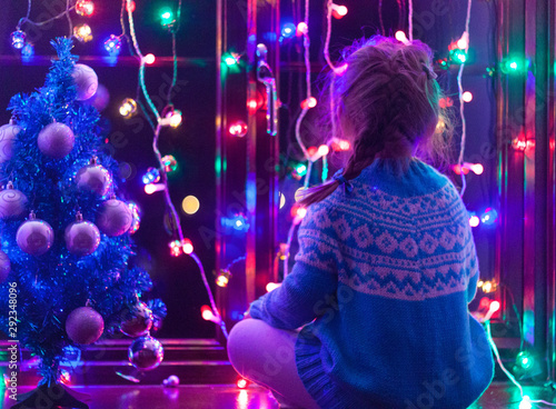 Happy little cute, beautiful girl 5 years old is sitting on the window and playing with Christmas lights and  Christmas decoration. Dark-blue theme. 