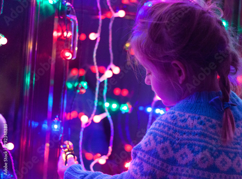 Happy little cute, beautiful girl 5 years old is sitting on the window and playing with Christmas lights and Christmas decoration. Dark-blue theme. 
