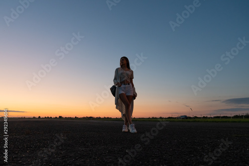 Young girl in white shorts and sneakers standing on the road against sunset