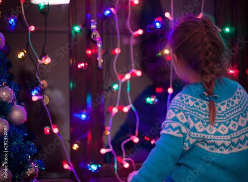 Happy little cute, beautiful girl 5 years old is sitting on the window and playing with Christmas lights and Christmas decoration. Dark-blue theme. 