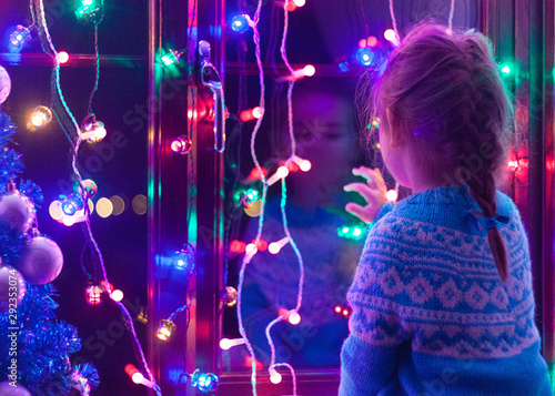 Happy little cute, beautiful girl 5 years old is sitting on the window and playing with Christmas lights and  Christmas decoration. Dark-blue theme. 