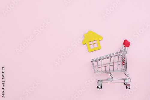 Shopping cart with a house on a pink background, top view. Place for text. Buying a home. Mortgage and loan. Concept of buying a house or apartment. Affordable housing. 