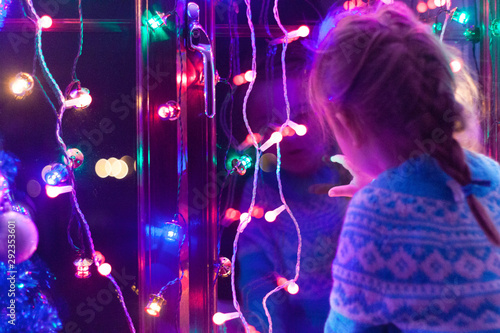 Happy little cute, beautiful girl 5 years old is sitting on the window and playing with Christmas lights and  Christmas decoration. Dark-blue theme. 
