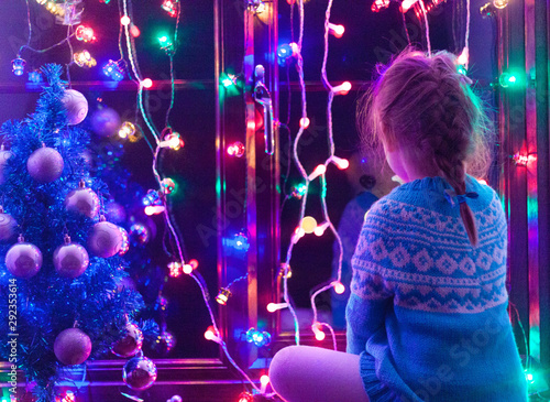 Happy little cute, beautiful girl 5 years old is sitting on the window and playing with Christmas lights and  Christmas decoration. Dark-blue theme. 