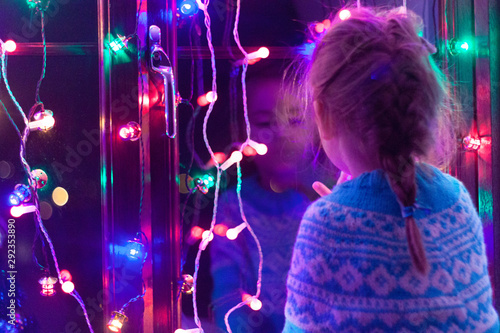 Happy little cute, beautiful girl 5 years old is sitting on the window and playing with Christmas lights and  Christmas decoration. Dark-blue theme. 