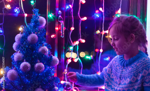 Happy little cute, beautiful girl 5 years old is sitting on the window and playing with Christmas lights and  Christmas decoration. Dark-blue theme. 