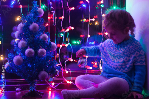 Happy little cute, beautiful girl 5 years old is sitting on the window and playing with Christmas lights and  Christmas decoration. Dark-blue theme. 