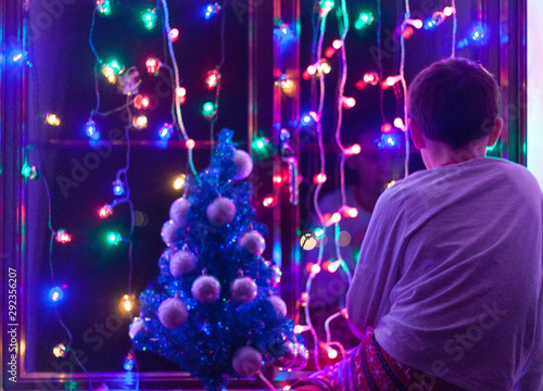 Teenager boy is sitting on the window and looking at the Christmas lights