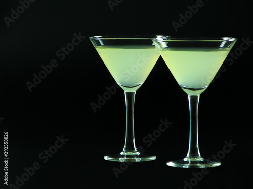 A classic daiquiri alcoholic cocktail of light green color from white rum, simple syrup and lime juice, in two conical cocktail glasses on a dark background