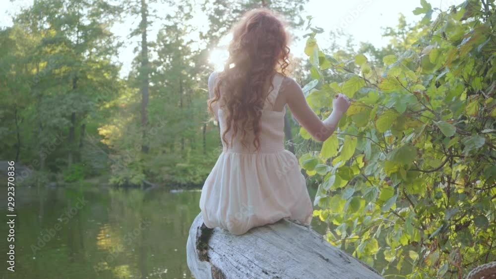 Back view of attractive slim caucasian woman with red hair sitting on the riverbank. Leisure outdoors. Connection with nature. Beautiful landscape. Rural life Back view of attractive slim caucasian woman with red hair sitting on the riverbank. Leisure outdoors. Connection with nature. Beautiful landscape. Rural life
