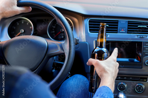 Closeup male hand is holding beer behind steering wheel of car. Drunk driver is riding in automobile. Man is violating rules of road. Alcohol driving concept. Deadly danger on route of modern city.