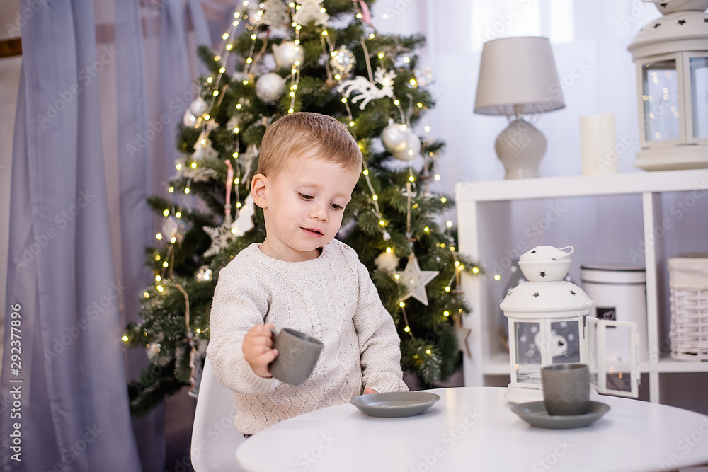 A little boy in a white pullover plays tea, drinks cocoa from a gray cup at his white table in the children's room. In the background there is a Christmas tree with garlands of lights. Copy space A little boy in a white pullover plays tea, drinks cocoa from a gray cup at his white table in the children's room. In the background there is a Christmas tree with garlands of lights. Copy space