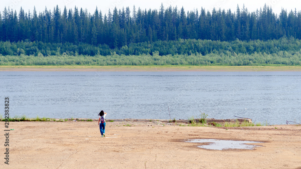 Φωτογραφία A tourist girl with a backpack touching her hair, lonely goes to the concrete cliff of the shore on the old pier at the Northern river vilyu with spruce tundra of the forest of Yakutia