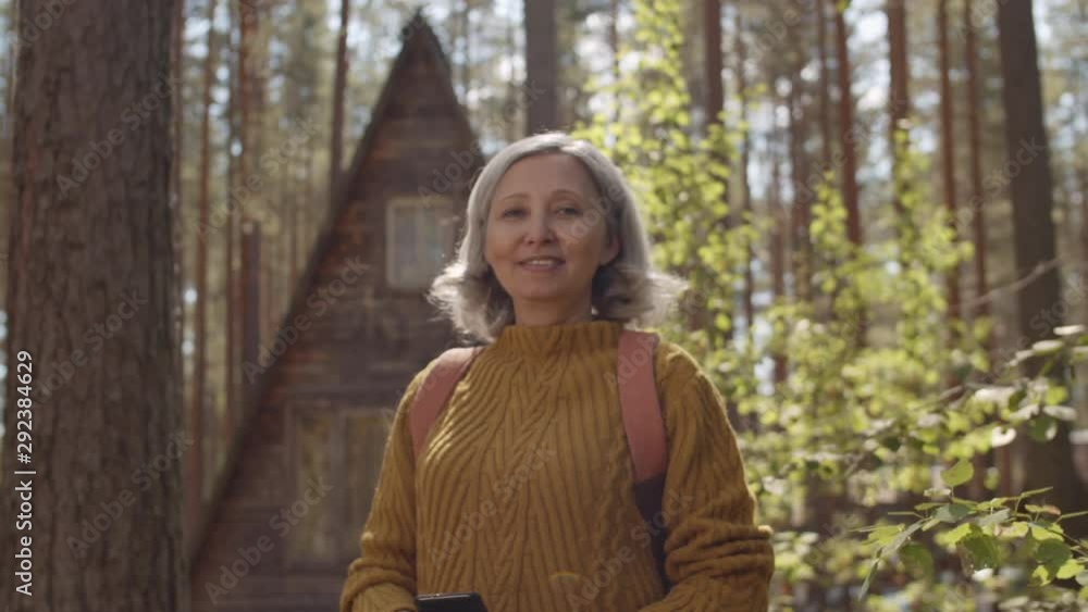 Medium shot of grey-haired female tourist wearing warm clothes and backpack standing in front of small wooden chalet in forest, looking aside and then looking at camera Medium shot of grey-haired female tourist wearing warm clothes and backpack standing in front of small wooden chalet in forest, looking aside and then looking at camera