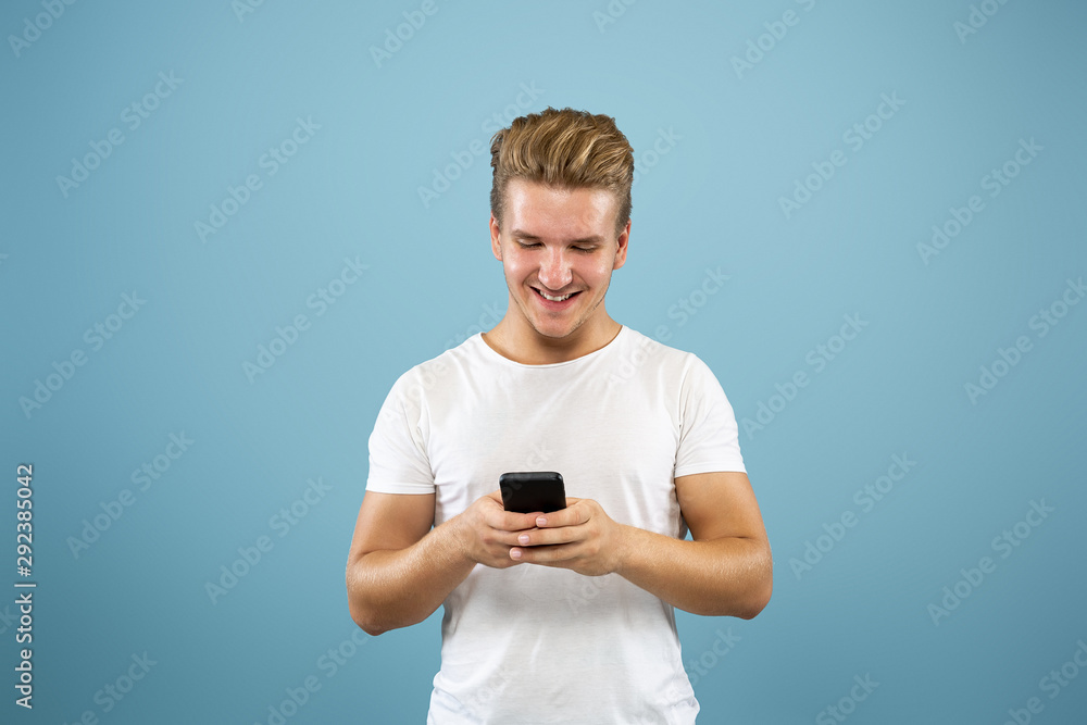 Caucasian young man's half-length portrait on blue studio background. Beautiful male model in shirt. Concept of human emotions, facial expression, sales, ad. Using phone, online payment, betting. Caucasian young man's half-length portrait on blue studio background. Beautiful male model in shirt. Concept of human emotions, facial expression, sales, ad. Using phone, online payment, betting.