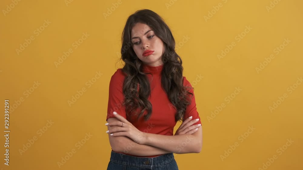 Casual bored brunette girl in red top with hands crossed tiredly looking around over yellow background. Not interested expression Casual bored brunette girl in red top with hands crossed tiredly looking around over yellow background. Not interested expression