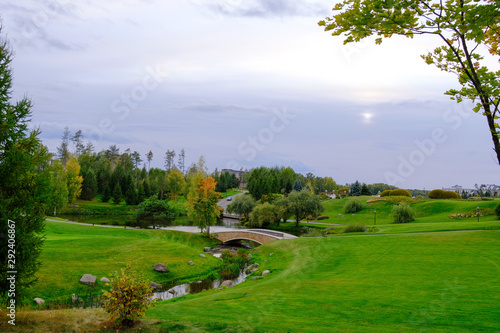 Autumn landscape. Hills with green lawn and ornamental shrubs and trees with a bridge over the creek against the evening sky. It's a beautiful park. © Александр Овсянников