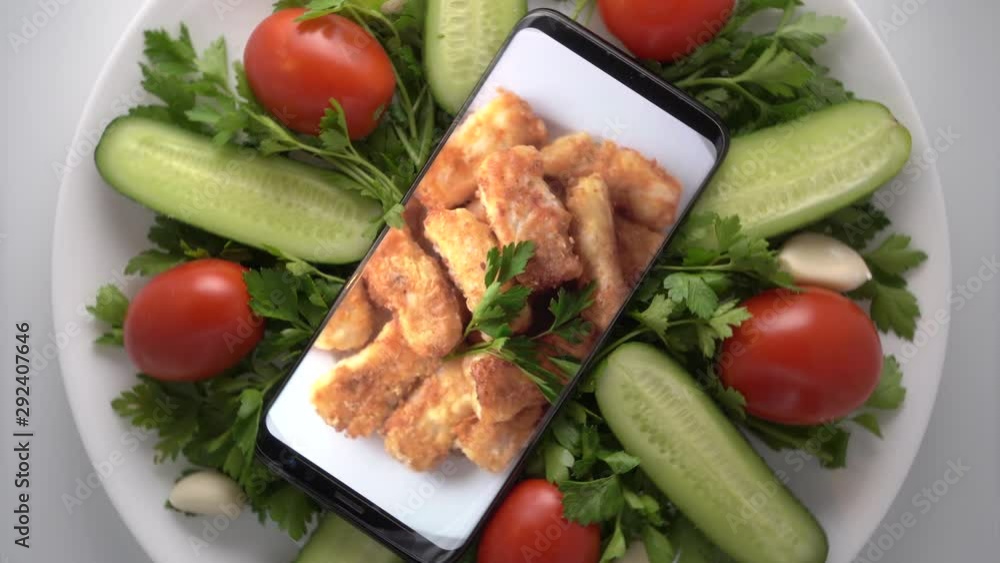 Plate with salad and cellphone with crispy fried cod fish fillet on screen. Food blog concept. Top view of mobile phone with food footage on screen rotating. Plate with salad and cellphone with crispy fried cod fish fillet on screen. Food blog concept. Top view of mobile phone with food footage on screen rotating.