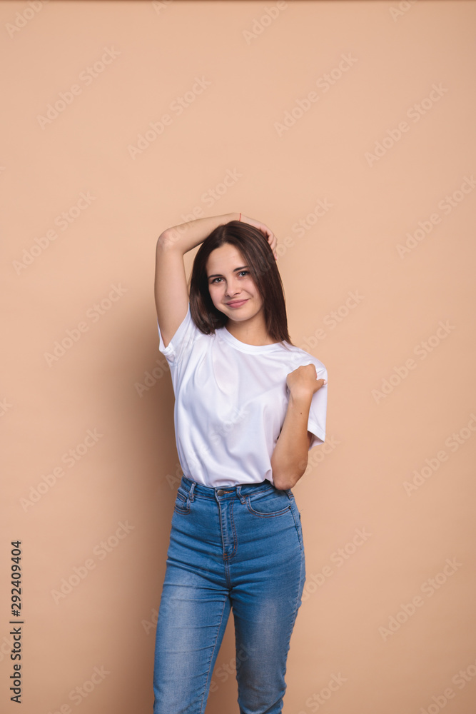 A girl looks at the camera and smiles, on a beige background, space for text. Portrait of a cute girl with clean skin who straightens her hair with her hands. Stylish girl in jeans and a white jacket A girl looks at the camera and smiles, on a beige background, space for text. Portrait of a cute girl with clean skin who straightens her hair with her hands. Stylish girl in jeans and a white jacket