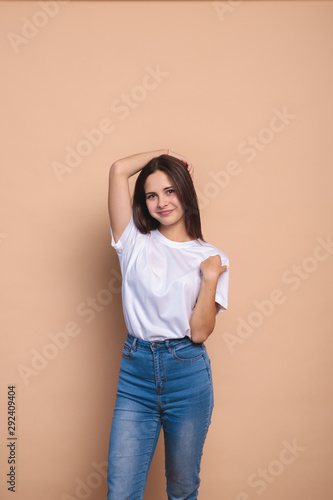 A girl looks at the camera and smiles, on a beige background, space for text. Portrait of a cute girl with clean skin who straightens her hair with her hands. Stylish girl in jeans and a white jacket