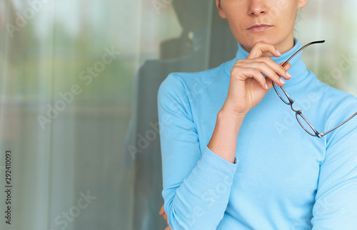 Horizontal cropped image of beautiful blonde young serious woman in office wearing blue turtleneck, holding eyeglasses in the hand. People and business concept